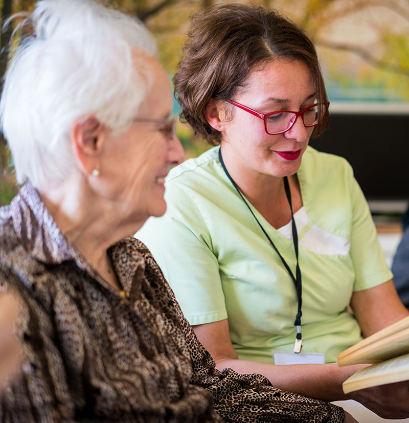 An elderly woman with white hair smiles while a carer in a green top reads to her.