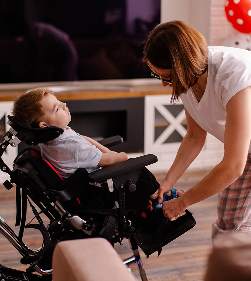 A caring adult secures a child's harness in a modern wheelchair within a cozy home setting.