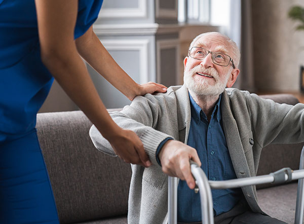 Elderly man with glasses smiling upwards at a caregiver in blue attire, holding onto a walking frame.