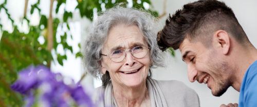 An elderly woman with glasses smiling broadly as a young man leans in close, sharing a joyful moment.