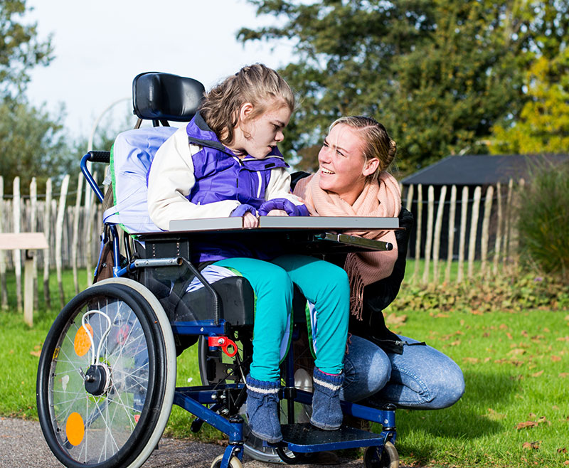 A young girl in a wheelchair sharing a joyful moment with her carer outdoors.