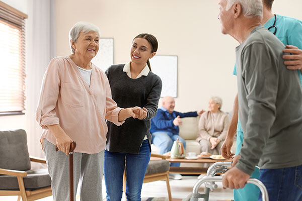 Elderly woman with a cane being assisted by a caregiver in a social care setting, with others in the background.