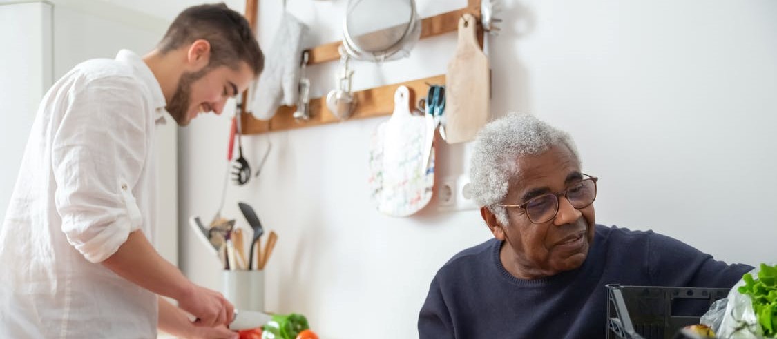 A young man preparing food in a kitchen while an older man watches.