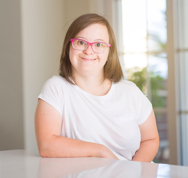 Smiling woman with Down syndrome wearing pink glasses, posing indoors.