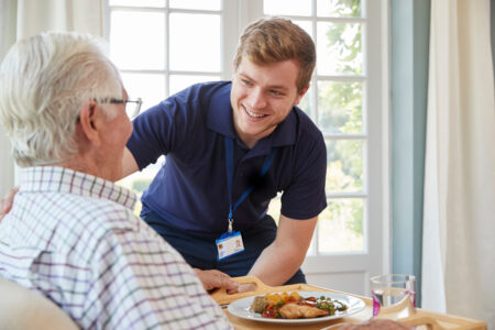 Young male caregiver serving meal to elderly person at home
