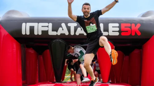 A man joyfully jumps above an inflatable archway at the finish line of a 5K run.