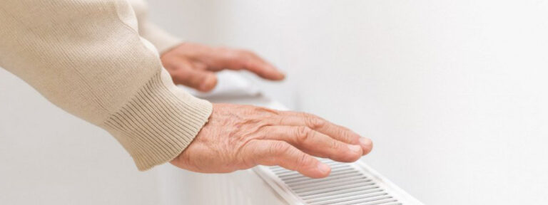 Close-up of an elderly person's hands warming up over a white radiator.