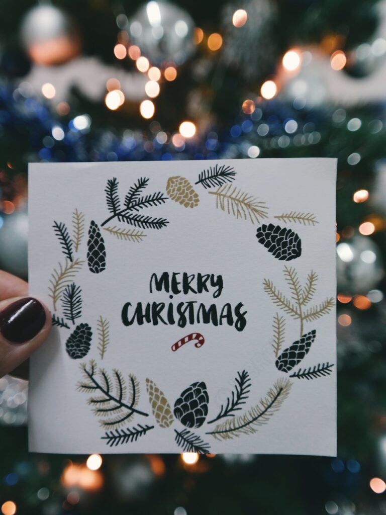 Hand holding a Christmas card with 'Merry Christmas' surrounded by pine illustrations, against a blurred tree backdrop.