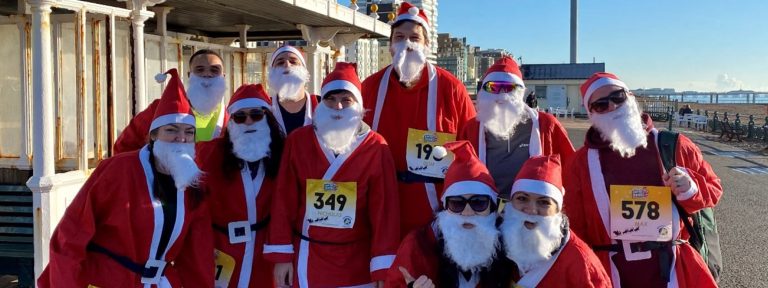 The image shows a group of people dressed in Father Christmas costumes, complete with red hats, jackets, and white beards. They are wearing numbers, suggesting they are participating in a themed fun run or charity race. They're posing for the photo with a backdrop of a classic seaside promenade, identifiable by the white balustrade and shelter, under a clear blue sky. The setting appears to be a bright, sunny day, likely along a coastal area, possibly during a festive season event.