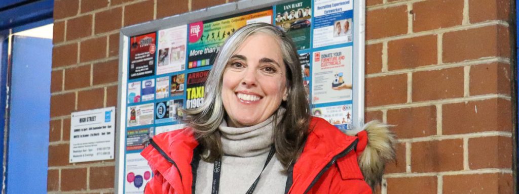 Smiling woman in a red jacket standing in front of a community noticeboard.