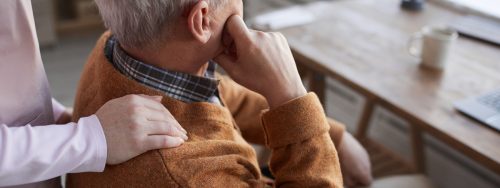 A supportive hand rests on the shoulder of an elderly man in thought, symbolizing care and comfort.