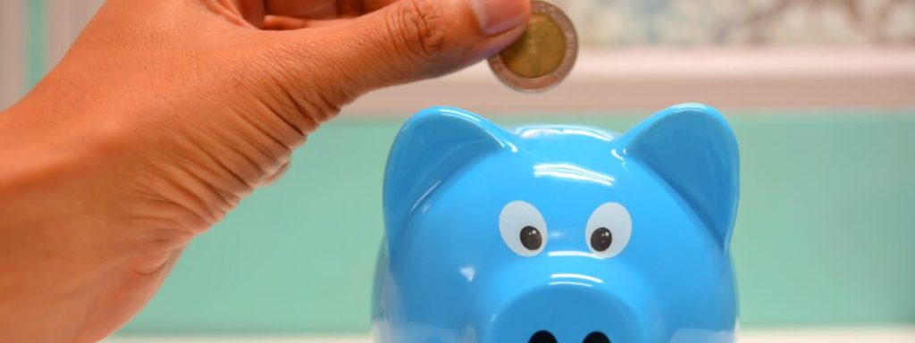 Close-up of a person's hand inserting a coin into a blue piggy bank.