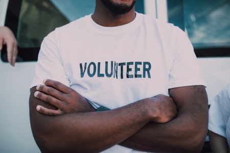 Close-up of a person with crossed arms wearing a white t-shirt with the word "VOLUNTEER" printed across the chest.