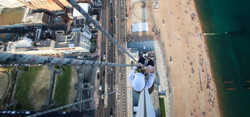 A bird's-eye view of an individual rappelling down a high building with a coastal road and beach visible below.