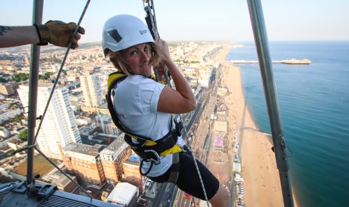 A woman smiles while rappelling down a seaside building, with a coastal cityscape stretching out below.