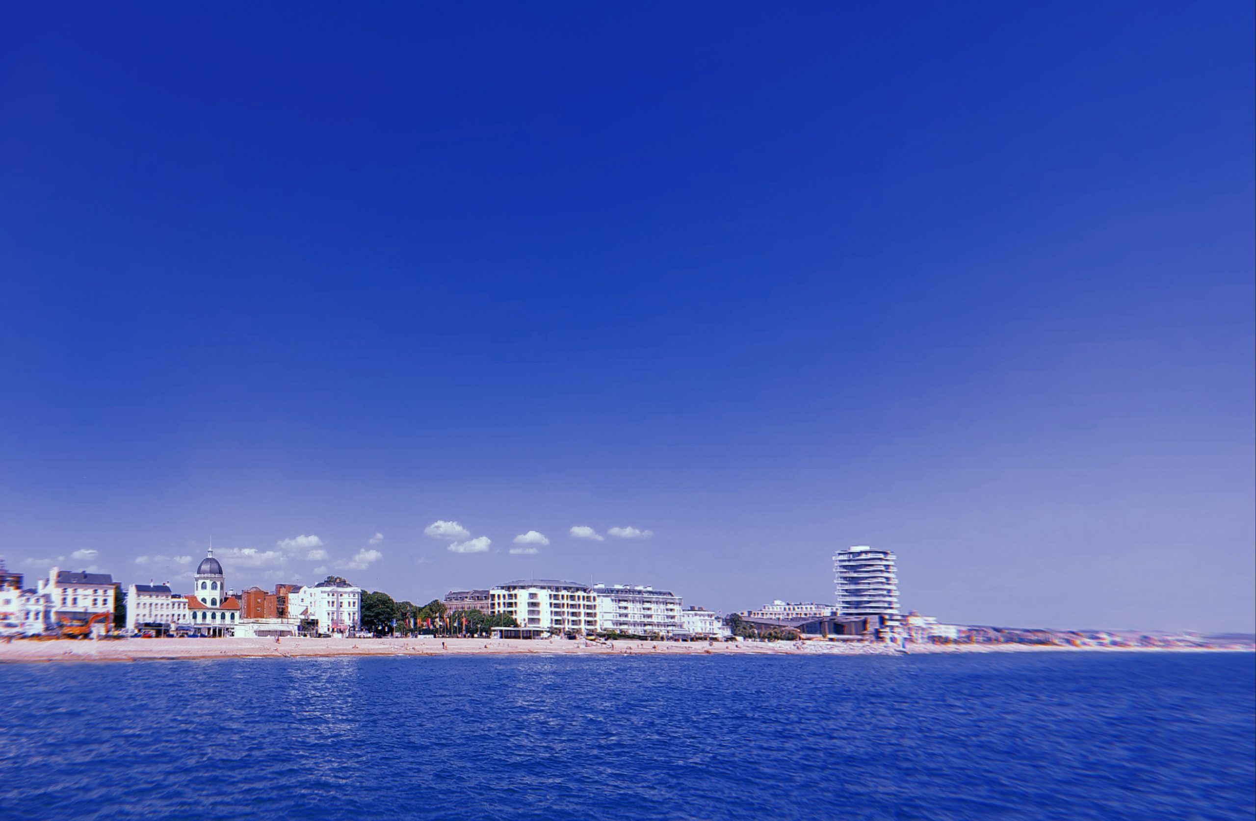 A serene seascape with a clear blue sky above and calm waters below. The coastline is dotted with buildings, including a distinctive structure with a domed roof.