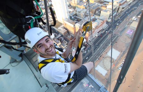 A young man with a beaming smile prepares to rappel down a building, with a bustling city street visible below.