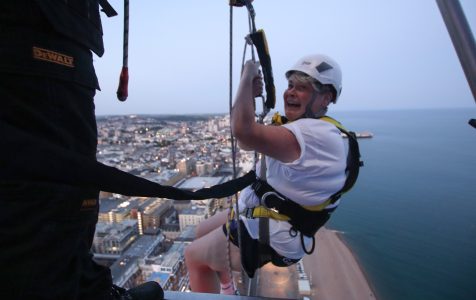A woman in a harness and helmet looks joyfully at the camera while rappelling down a high building with a city and coastline in the background.