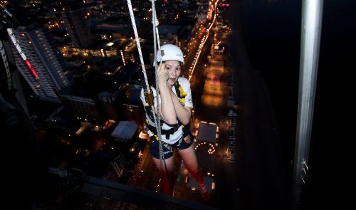 A woman begins her descent rappelling down a high building at night with city lights below.