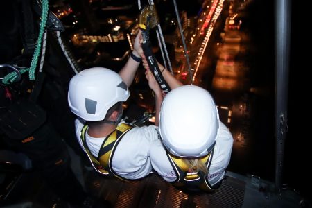 Two people in white helmets and harnesses preparing to rappel at night above city lights.
