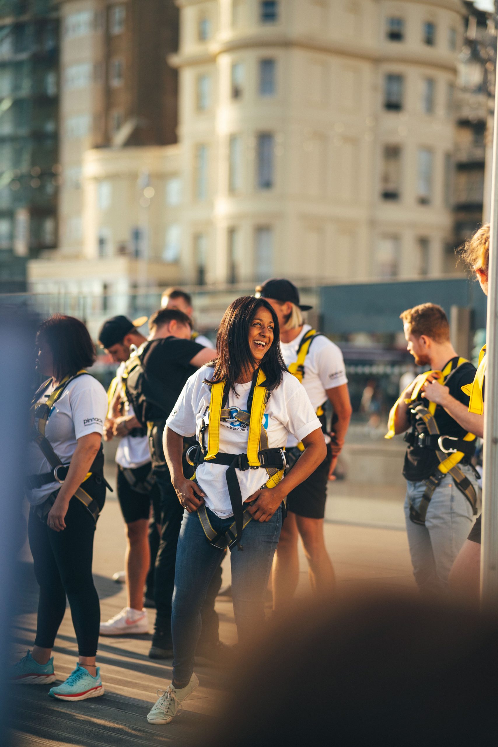 A smiling woman in a harness stands among a group of people preparing for an adventurous activity.