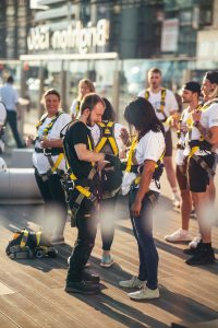A group of people in safety harnesses preparing for an activity, with a focus on a man and woman in conversation.
