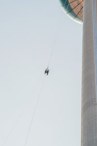 A person rappelling down a high structure with a large observation deck at the top against a bright sky.