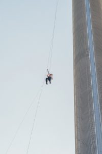 An individual in a red helmet is abseiling down a tall building at dusk.