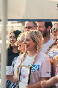 A smiling woman with braided hair, wearing sunglasses and a t-shirt with "Drop 360 Supporting Independent Lives" logo, holding a mug and a certificate, with a group of happy people in the background.