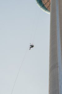 An individual in a harness is abseiling down a tall, modern building, with clear skies in the background.