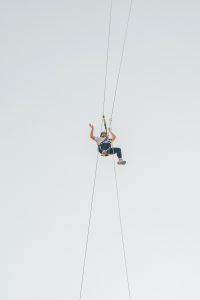 A person suspended in mid-air while abseiling down a building, with arms outstretched and a clear sky in the background.