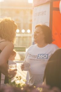 A joyful woman holding a mug converses at a sunny outdoor event, wearing a "Drop360 Supporting Independent Lives" t-shirt.