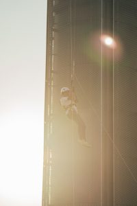 A person in safety gear is abseiling down a tall building, with the sun creating a halo effect around them.