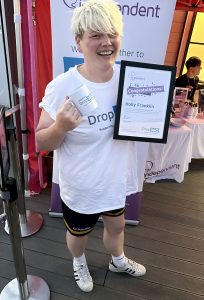 Beaming individual holding a certificate and a mug, standing in front of a banner for Independent Lives