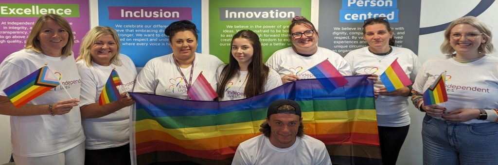 A group of individuals in white t-shirts with rainbow logos holding pride flags, standing in front of a banner with the words 'Excellence', 'Inclusion', 'Innovation', and 'Person centred'.