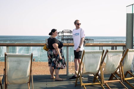 Two Independent Lives volunteers having a conversation on a seaside deck with a clear view of the ocean and a pier in the background.