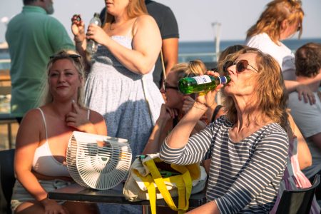 A woman in sunglasses enjoys a refreshing drink from a green bottle at a sunlit outdoor gathering.