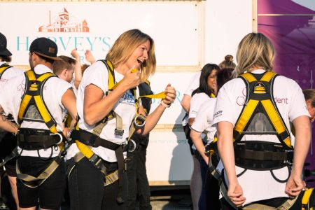 A woman in a white t-shirt and safety harness smiles brightly during a group outdoor activity.