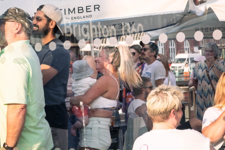 People gathered for a seaside event under a "Nyetimber" parasol, laughing and socialising.