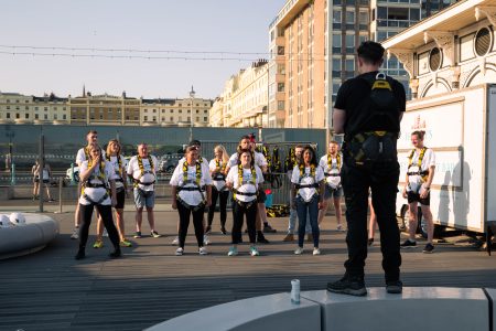 A group of focused abseiling participants in safety gear listen to an instructor during a briefing at an outdoor charity event.