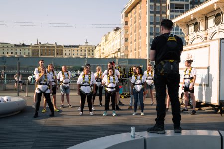 A group of focused abseiling participants in safety gear listen to an instructor during a briefing at an outdoor charity event.