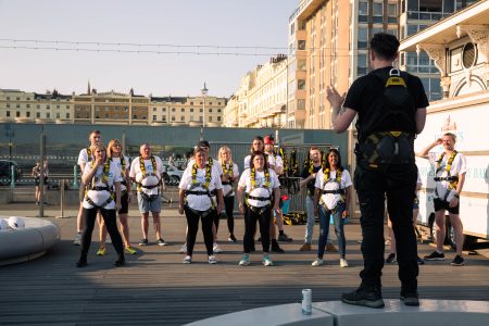 A group of focused abseiling participants in safety gear listen to an instructor during a briefing at an outdoor charity event.