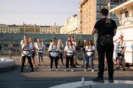 A group of focused abseiling participants in safety gear listen to an instructor during a briefing at an outdoor charity event.