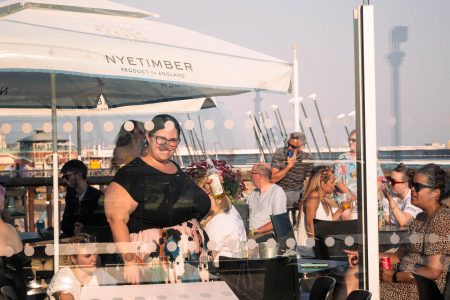 A woman laughs joyfully at an outdoor event under a parasol, with guests enjoying the seaside ambiance.