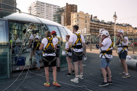 Abseiling participants prepare for a charity event on an outdoor platform with urban buildings in the background.