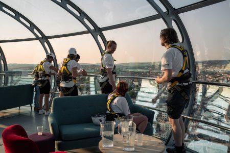 Abseil participants with safety gear enjoy the view from a glass-enclosed structure before their charity event.