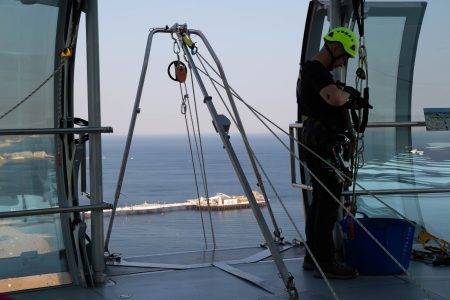 An abseiling instructor checks equipment on a high-altitude platform overlooking the ocean.