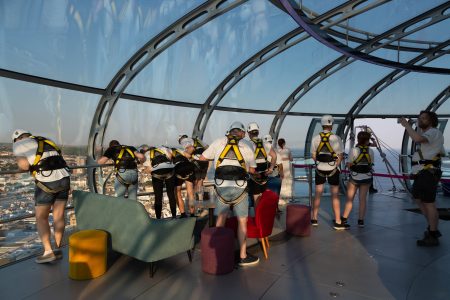 Participants in a charity abseil event prepare at a modern glass venue with a cityscape in the background.