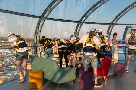 Participants in a charity abseil event prepare at a modern glass venue with a cityscape in the background.