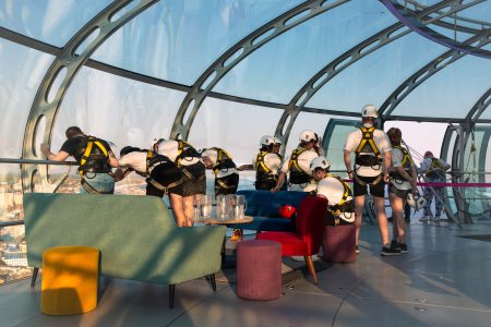 Participants in a charity abseil event prepare at a modern glass venue with a cityscape in the background.